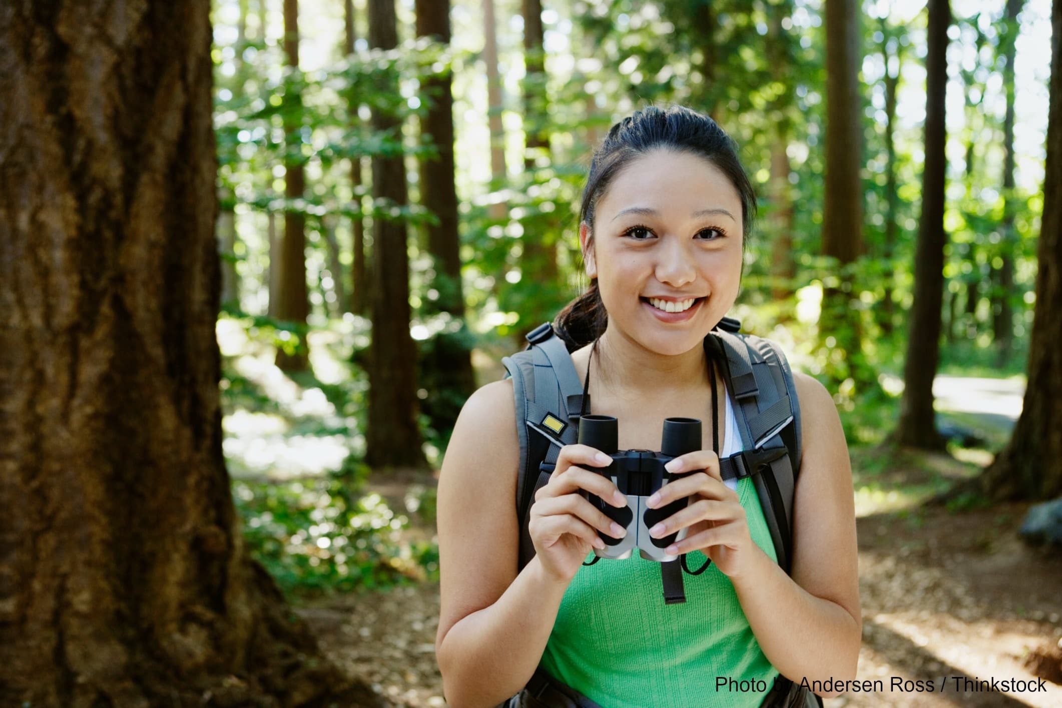 A smiling young woman holds binoculars while standing in a forest.