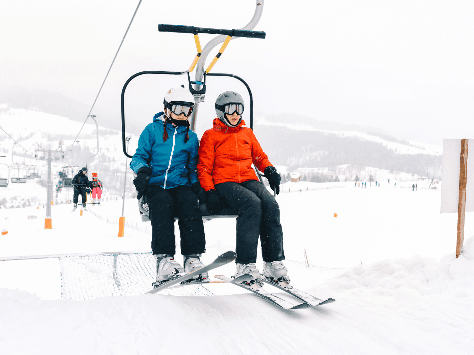 Two skiers wearing helmets and colorful jackets ride a ski lift in a snowy landscape.