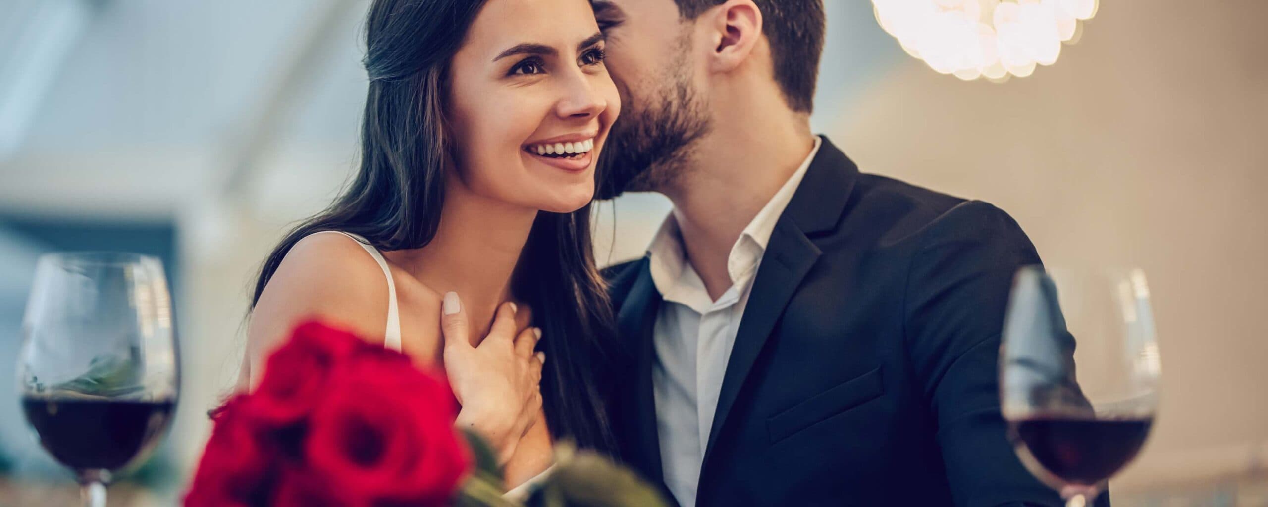 A smiling couple shares an intimate moment at a romantic dinner with roses and glasses of wine.
