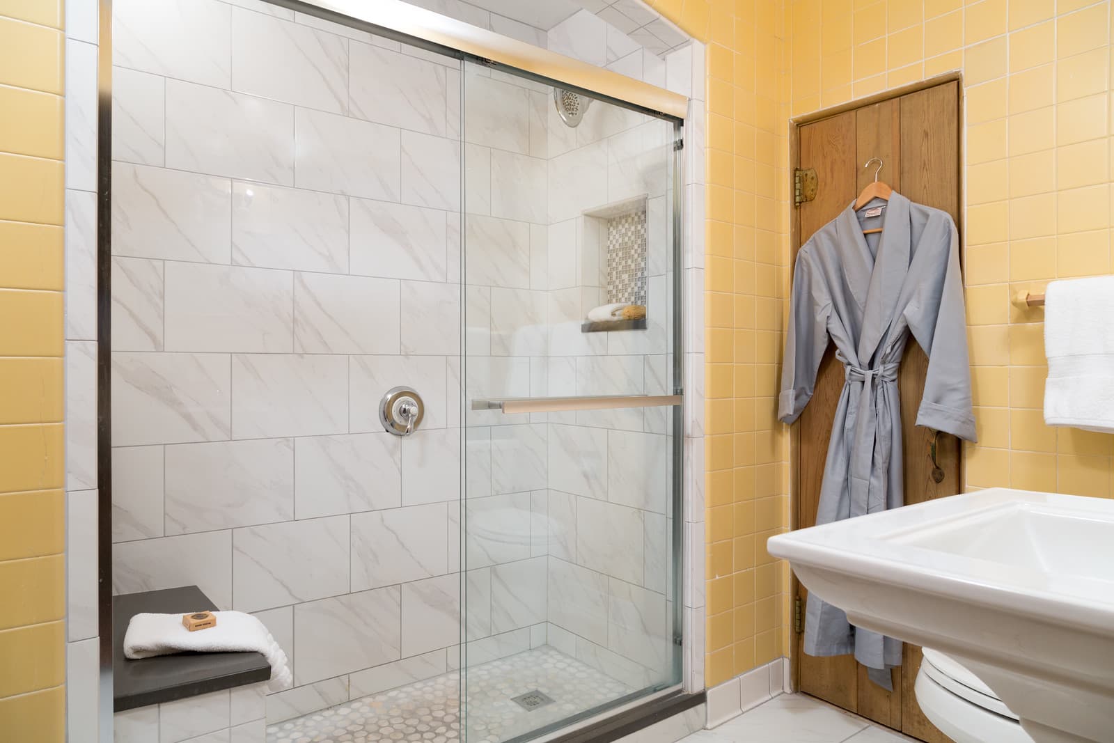 A modern bathroom featuring a glass shower, yellow tiled walls, and a bathrobe hanging on the door.