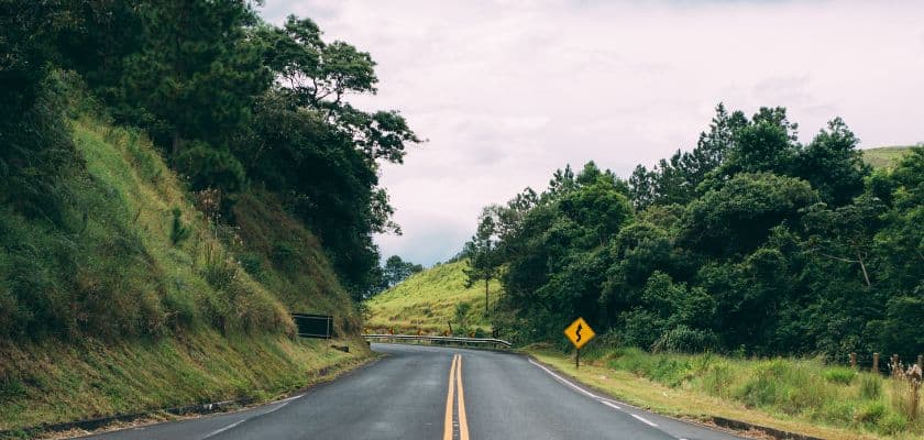 A picturesque winding road surrounded by lush greenery and a caution sign ahead.