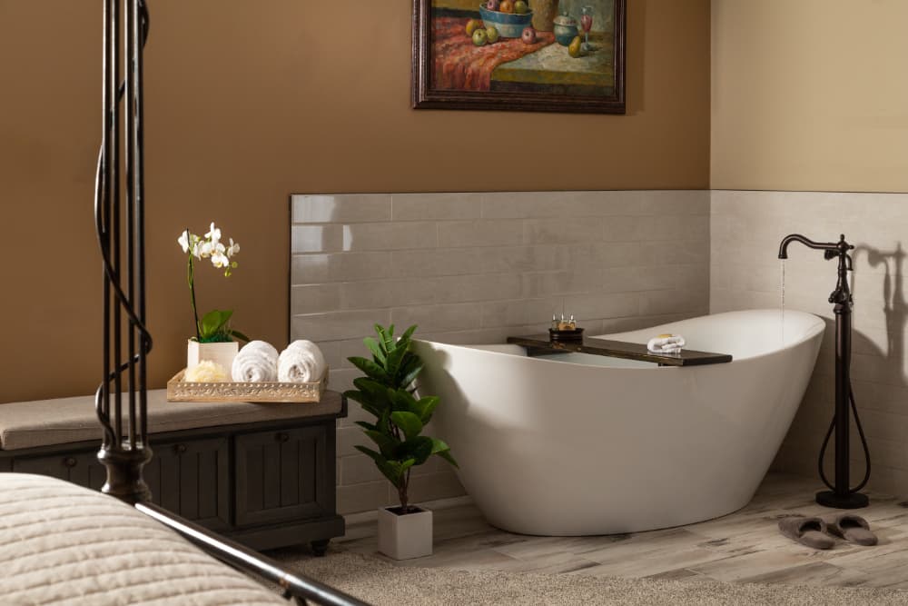 A luxurious freestanding bathtub in a modern bathroom. The bathtub is white and features a wooden tray with bath products. A large painting hangs on the wall behind the tub, and a potted plant sits on the floor next to it. A bed with a dark wooden headboard is visible in the background.