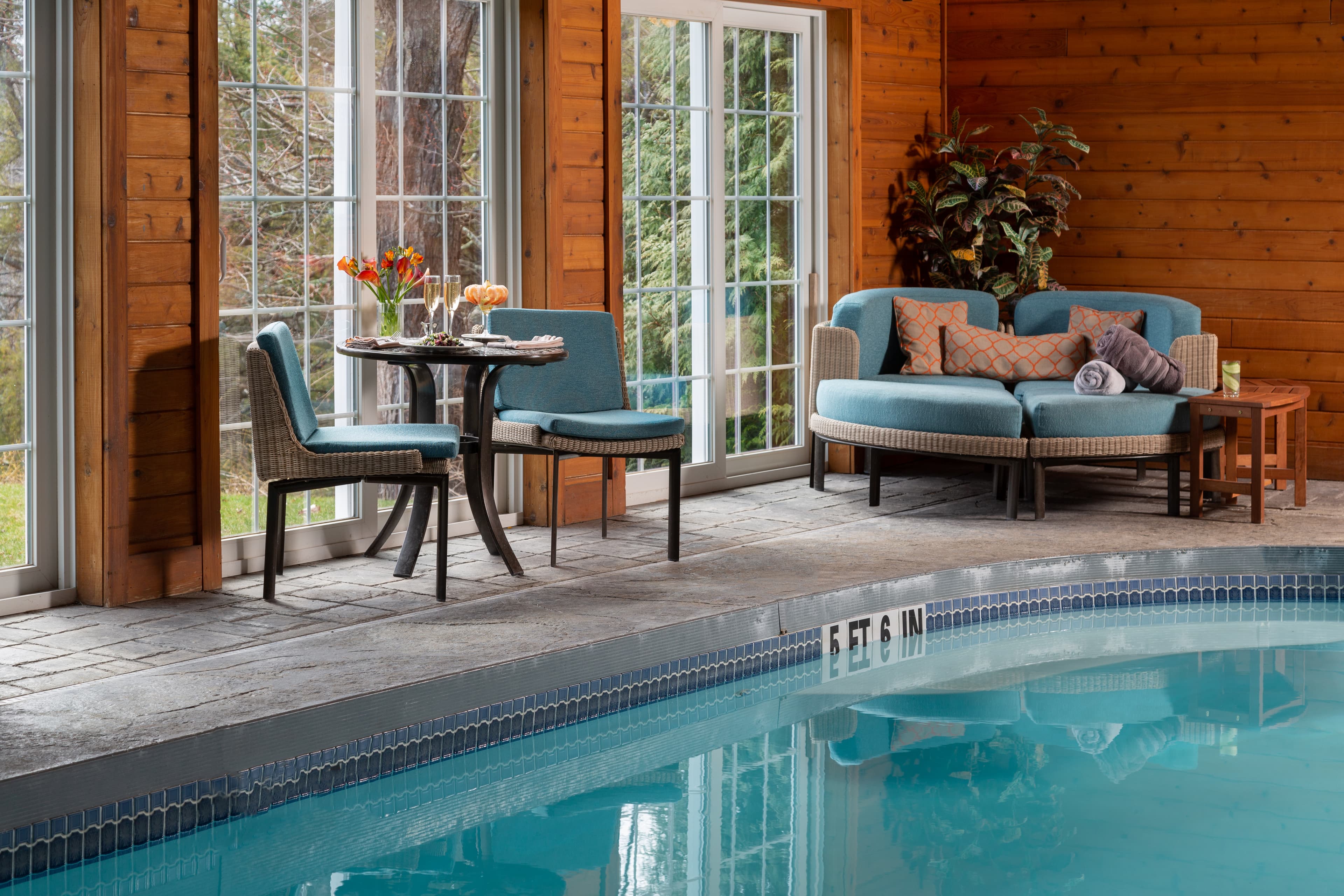 An indoor pool area with large windows overlooking a lush green landscape. The pool is surrounded by comfortable seating, including a small table with chairs and a curved sofa with pillows. The ceiling is high with exposed wooden beams.