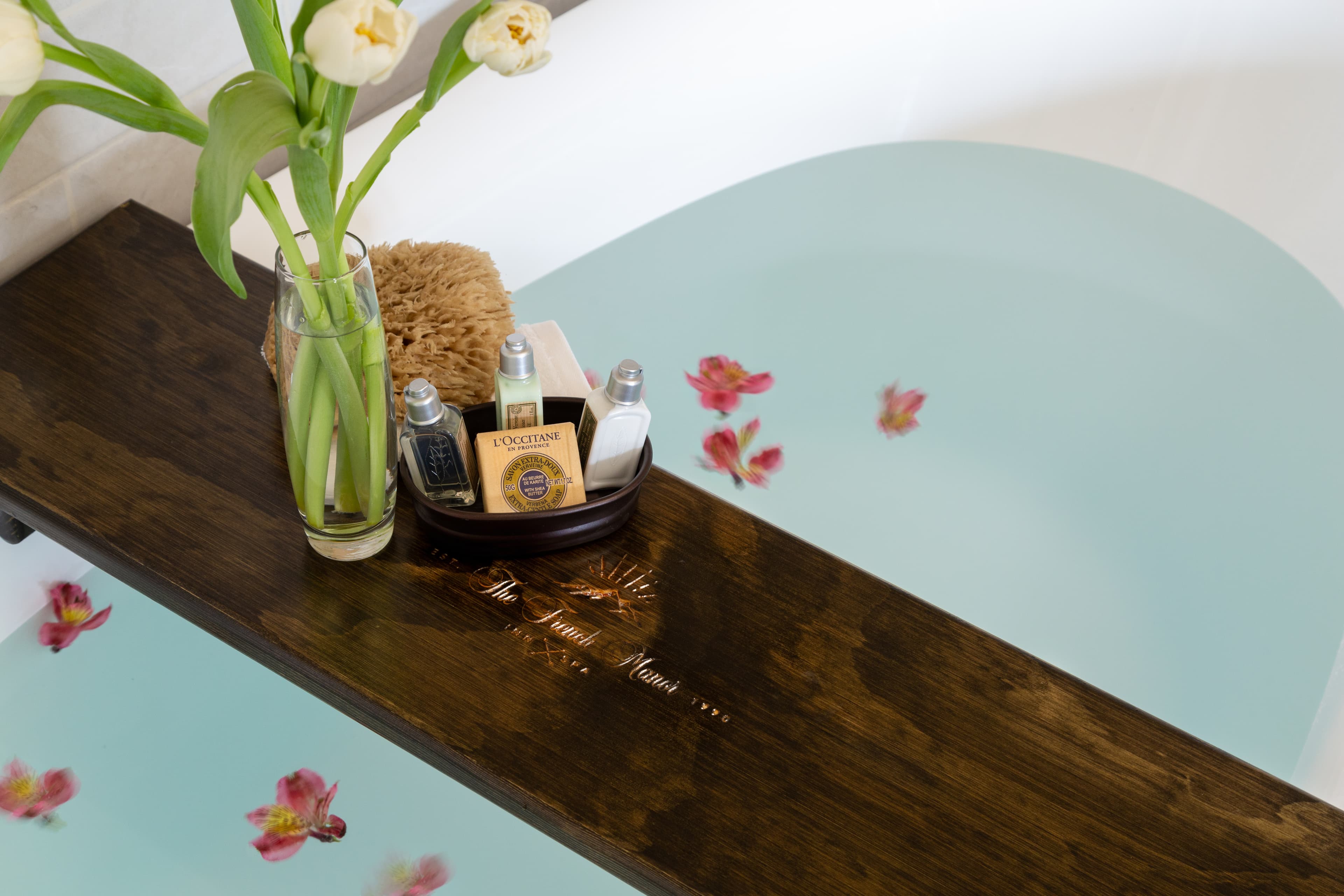 A wooden bath tray holds toiletries and a vase of flowers above a tranquil, water-filled bathtub adorned with floating petals.
