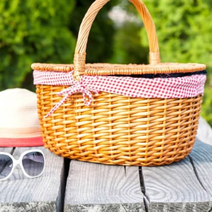 A wicker picnic basket with a red and white checkered lid, a straw hat, and sunglasses on a wooden table.