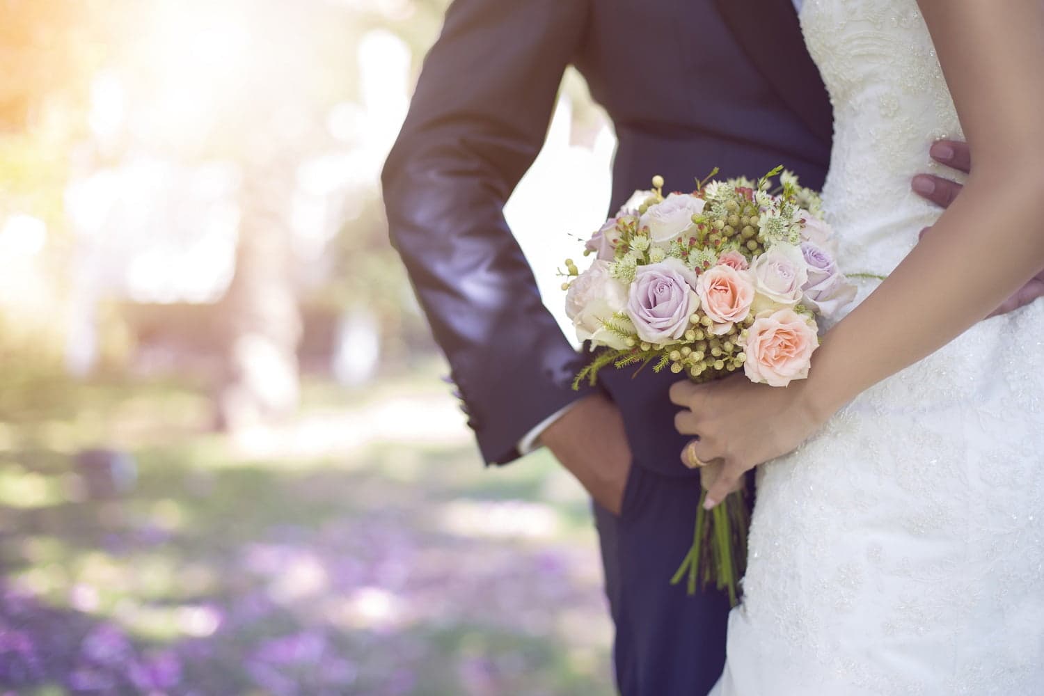 A bride in a white dress holds a floral bouquet while standing next to a groom in a navy suit, set in a softly lit outdoor setting.