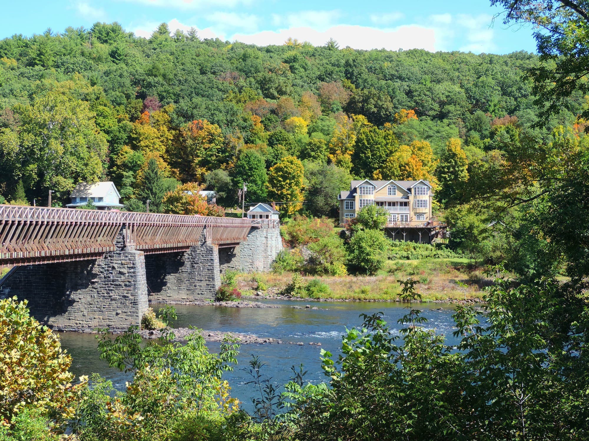 A scenic view of a wooden bridge crossing a river, with a house and colorful autumn trees in the background.
