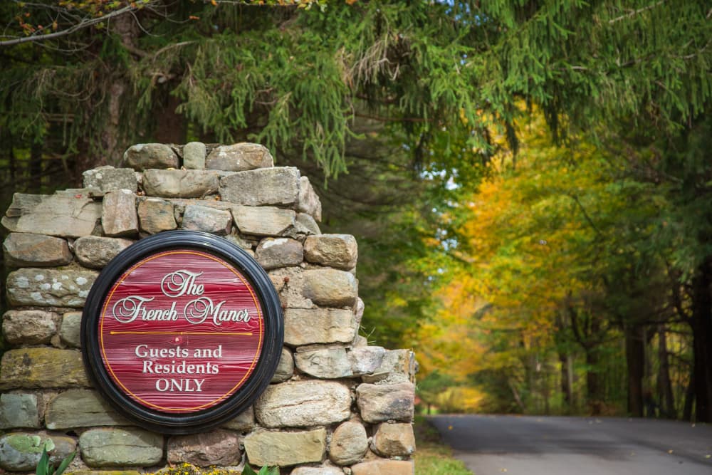 A stone entranceway to The French Manor with a circular sign featuring the establishment's name and the phrase "Guests and Residents Only." The sign is surrounded by lush greenery and colorful autumn foliage.