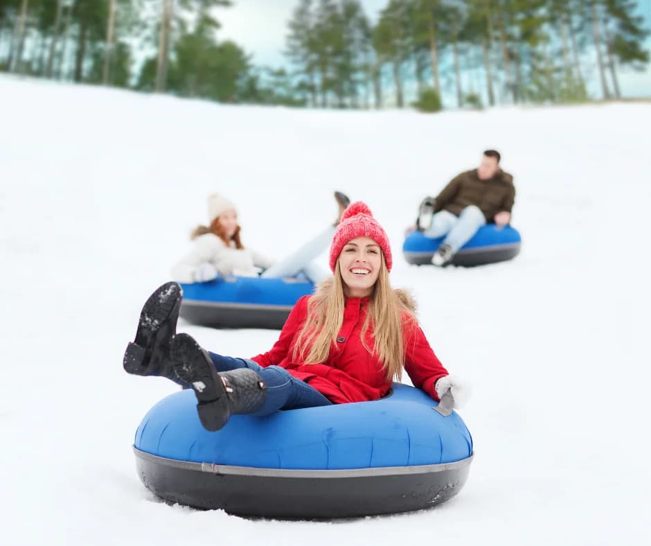 A smiling woman in a red jacket and pink hat enjoys snow tubing with friends in the background.