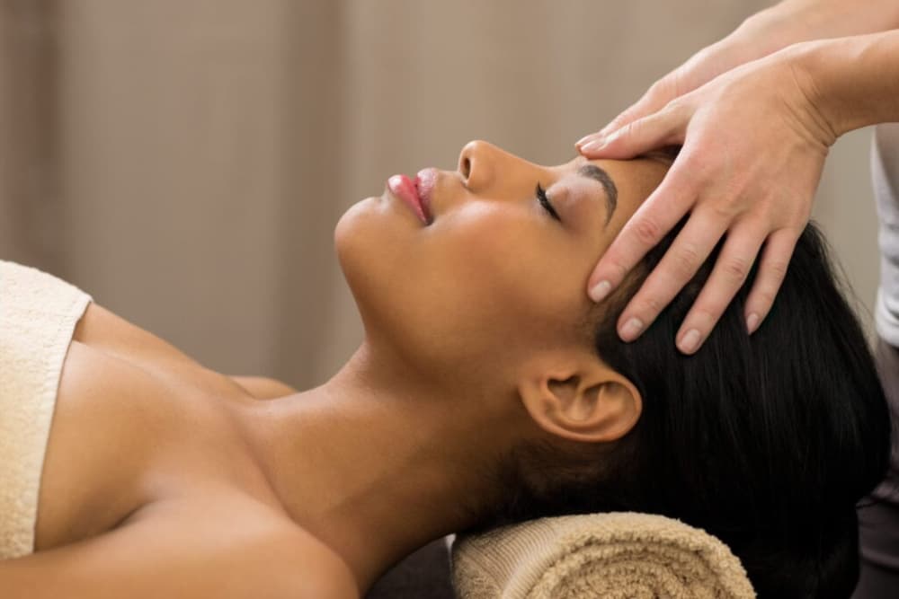 A woman laying face on on a table with a tan rolled towel under her neck, getting a head massage.