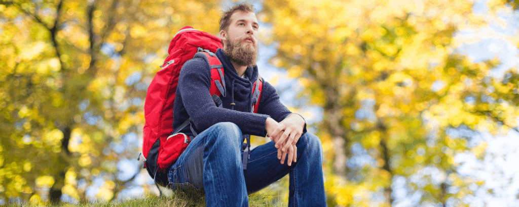 A bearded man sits on grass with a red backpack, surrounded by autumn foliage.
