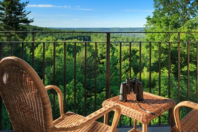 A balcony with a wicker chair and table, overlooking a lush green landscape with binoculars on the table.