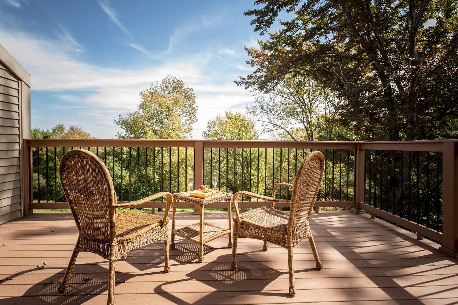 A cozy deck with two wicker chairs and a small table overlooking lush greenery under a clear blue sky.