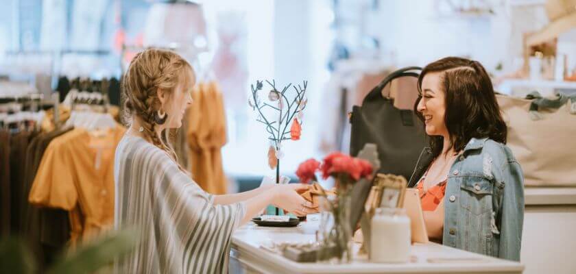 Two women interact in a boutique, with one handing an item to the other over a counter.