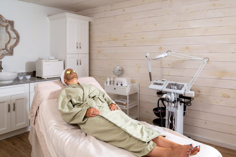 A woman laying on a massage table in a light green bathrobe with a mud mask on her face and sliced cucumbers over her eyes.