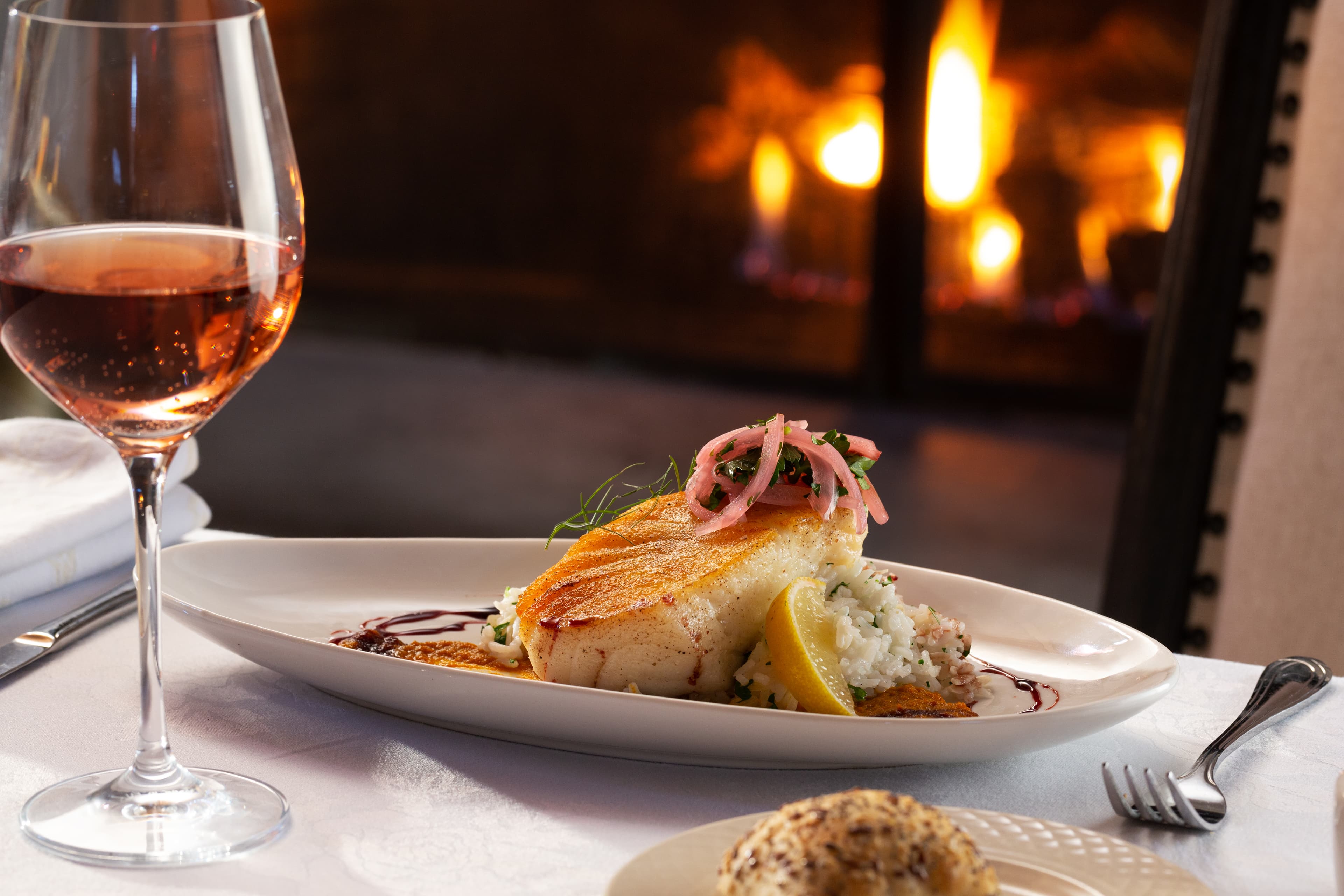 A plate of seared halibut over a delicate sauce, served with a bed of risotto and topped with pickled onions. A glass of rosé wine is placed next to the plate, and a fireplace is visible in the background.