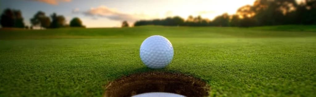 A golf ball resting on the lip of a golf hole, with a lush green golf course and a beautiful sunset in the background.