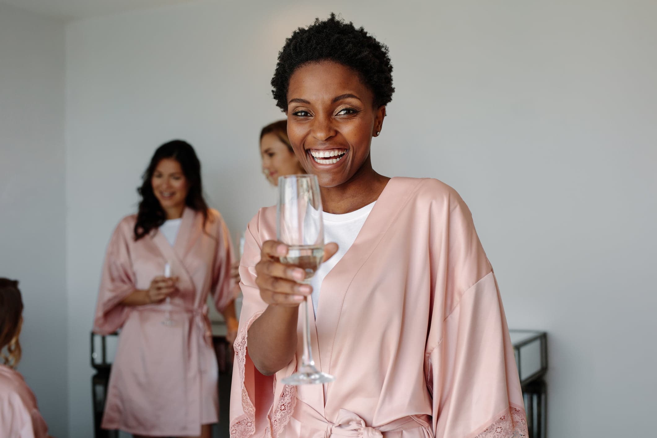 A smiling woman in a pink robe holds a champagne glass while celebrating with friends.