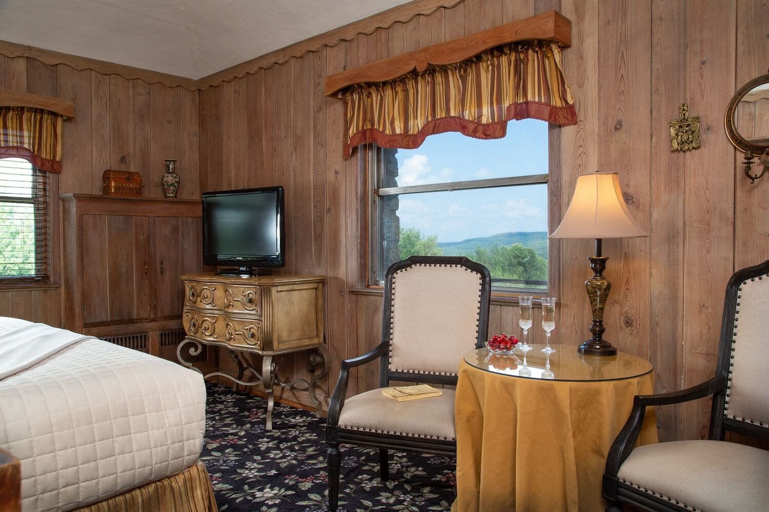 Cozy hotel room featuring wooden walls, a TV, and a small seating area with two chairs and a lamp.