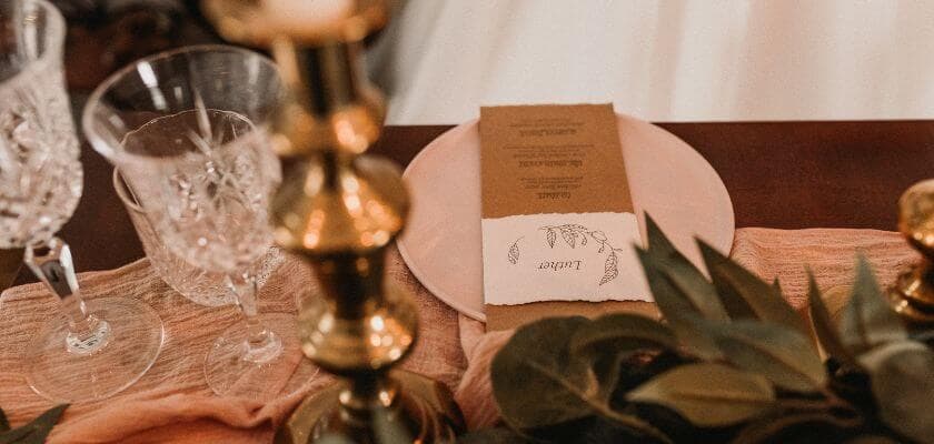A beautifully set dining table featuring crystal glasses, a candle holder, and a menu on a plate surrounded by greenery.