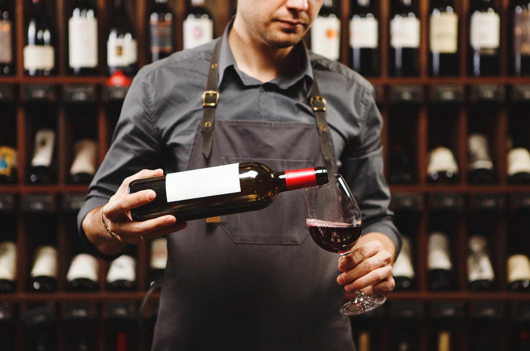A man in an apron pours red wine into a glass in front of a wine rack.