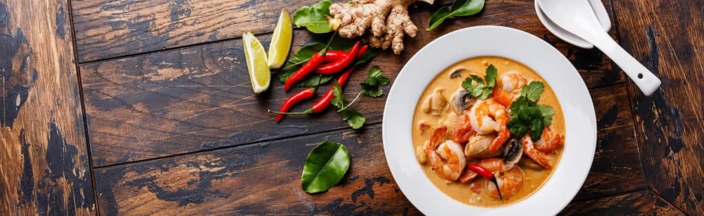 A bowl of Tom Yum soup with shrimp, mushrooms, lemongrass and chili peppers in a white bowl on a distressed wood table, along with an Asian soup spoon, along with pieces of fresh ginger, peppers, limes, and green leaves.
