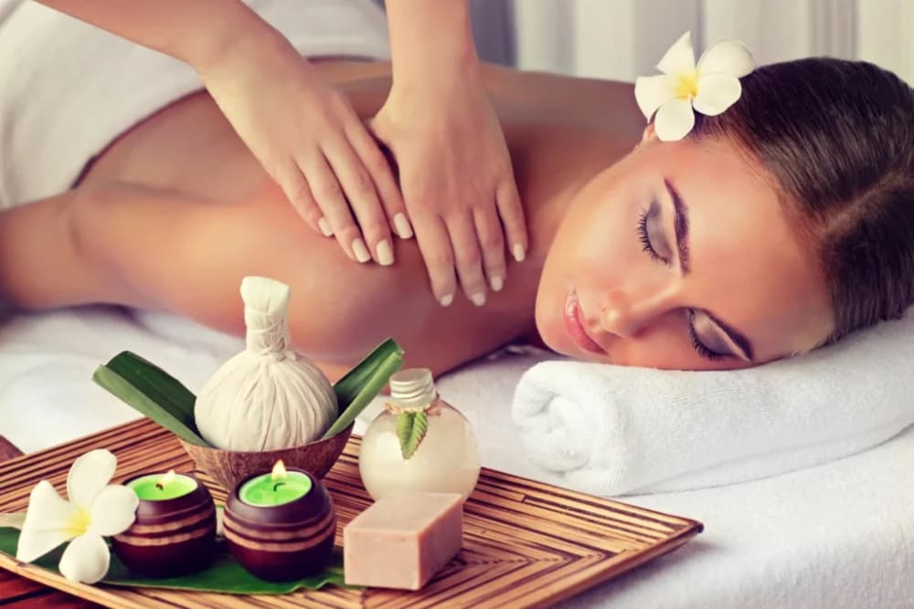 A woman laying face down on a massage table with a rolled towel under her face, a flower behind her ear, getting a back massage, along with a bamboo tray on the edge of the table with 2 tealight candles, spa amenities and a flower.