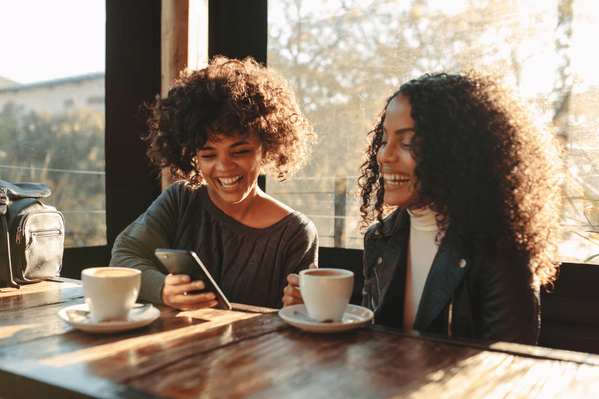 Two women with curly hair smile while sharing a moment over coffee at a cozy café.
