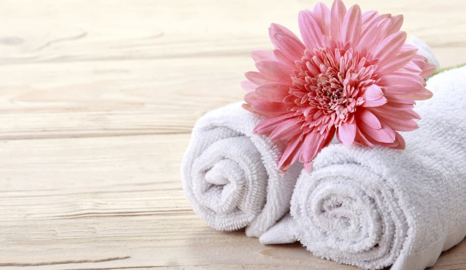 A wood table with 2 rolled white spa towels and a large pink flower on top of them.