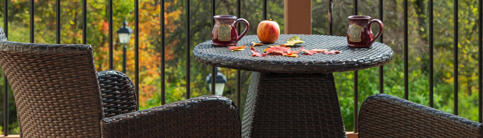 A cozy outdoor patio setting with two wicker chairs, a small round table, and two mugs filled with a warm beverage. An apple and fallen leaves are scattered on the table, and a scenic view of autumn foliage is visible in the background.