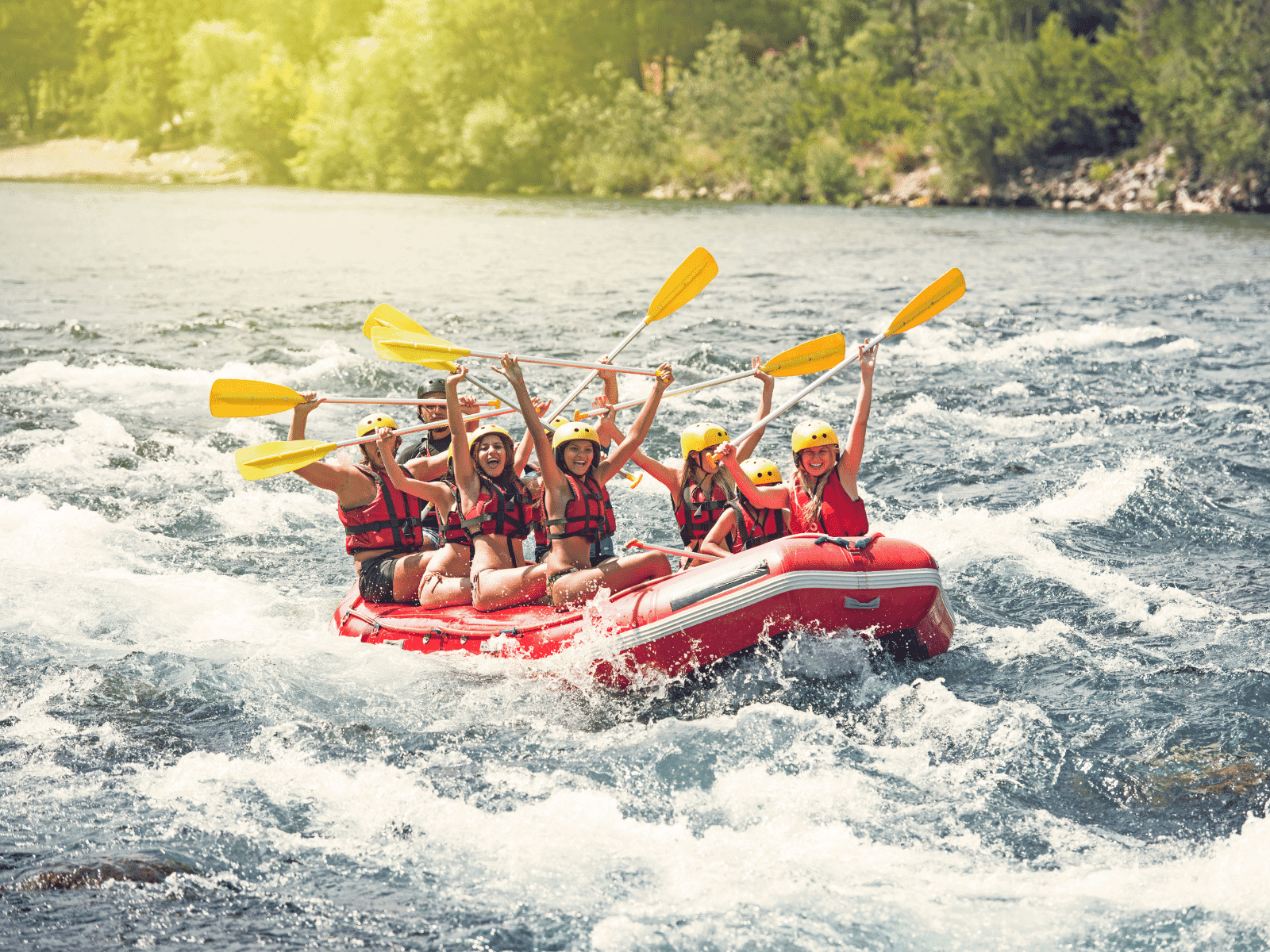 A group of excited individuals raft down a river, paddling with yellow oars and wearing life jackets and helmets.