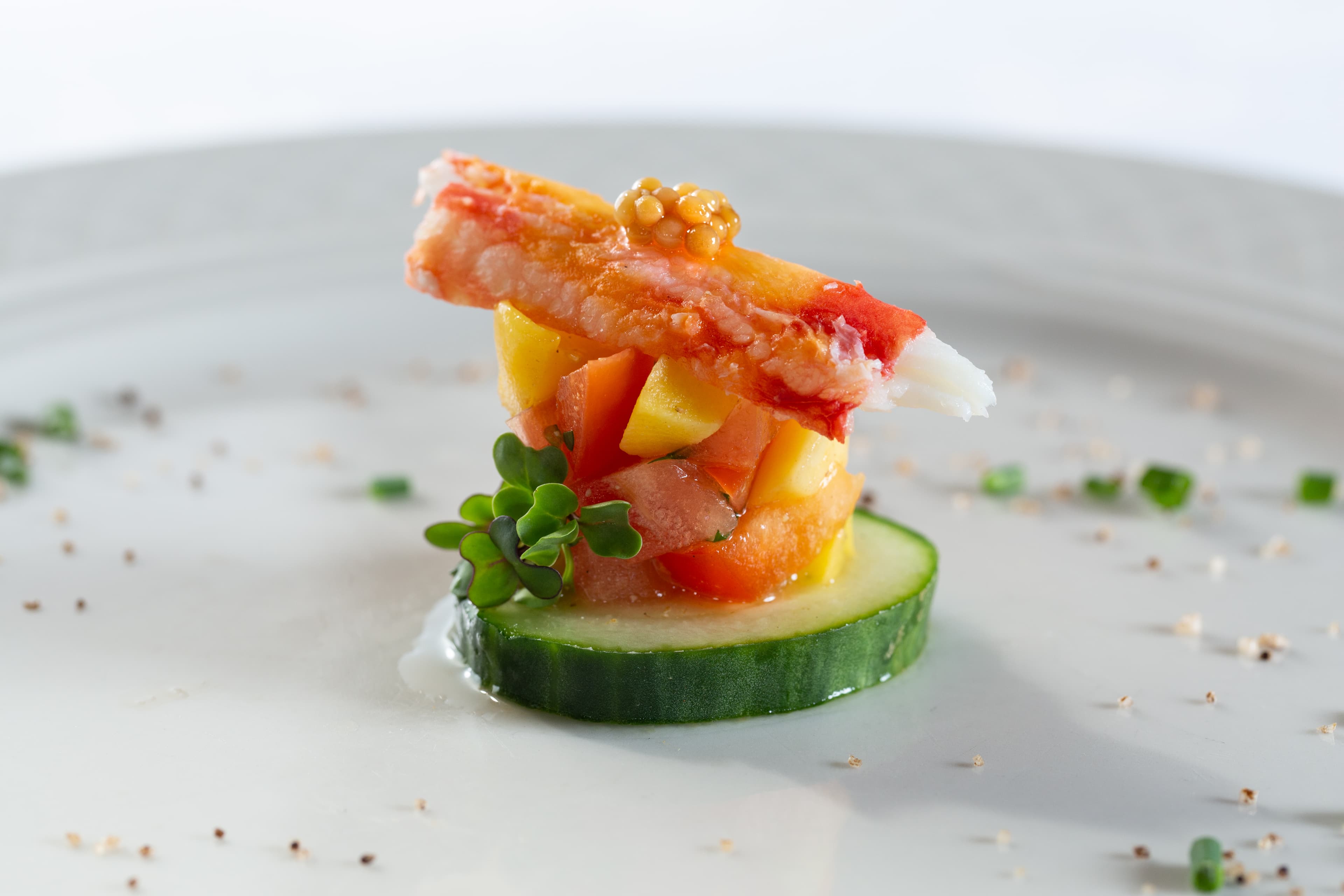 A small, elegant appetizer on a white plate. It features a crab claw, a layered tomato and cucumber salad, and a dollop of mustard. The dish is garnished with microgreens and chives.