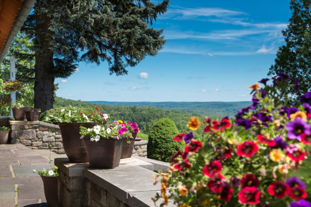 A scenic view from a stone patio with colorful flower pots. The patio overlooks the Poconos mountains and lush greenery under a clear blue sky.