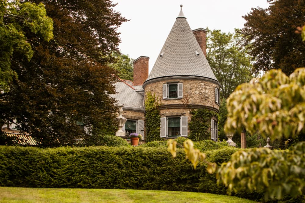 A historic stone mansion with a conical roof and ivy-covered walls. The building is surrounded by lush green trees and hedges.