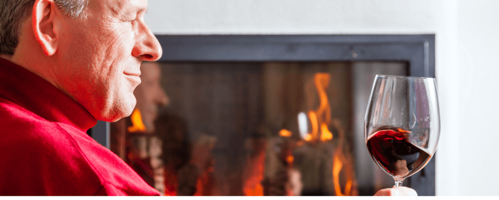 A man holds a glass of red wine while sitting beside a fireplace.