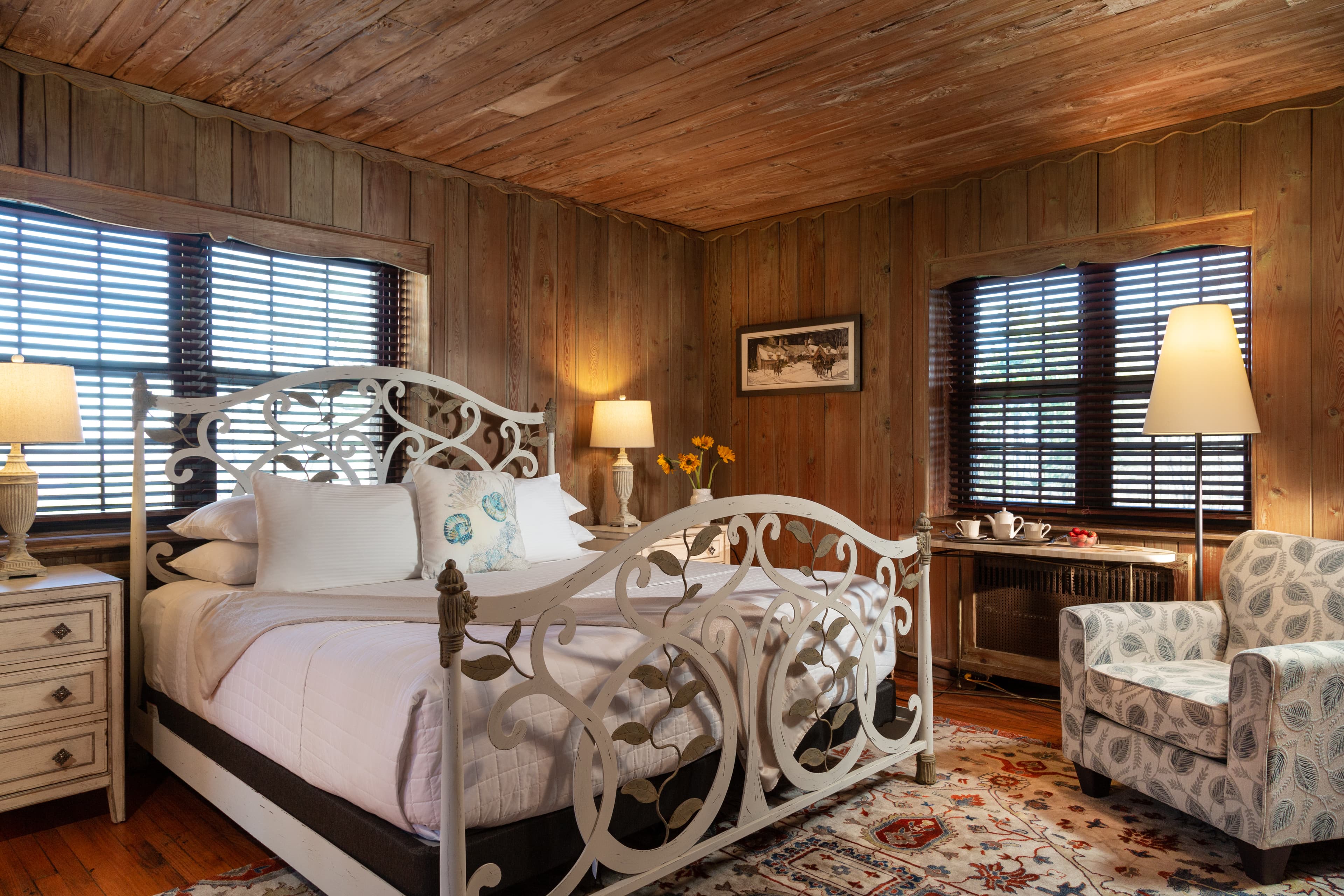 A cozy bedroom featuring a white ornate bed, bedside tables, a patterned armchair, and wooden walls with natural light streaming through windows.