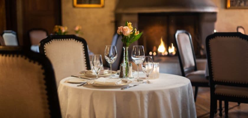 A elegantly set dining table with flowers and wine glasses in a cozy restaurant featuring a fireplace in the background.