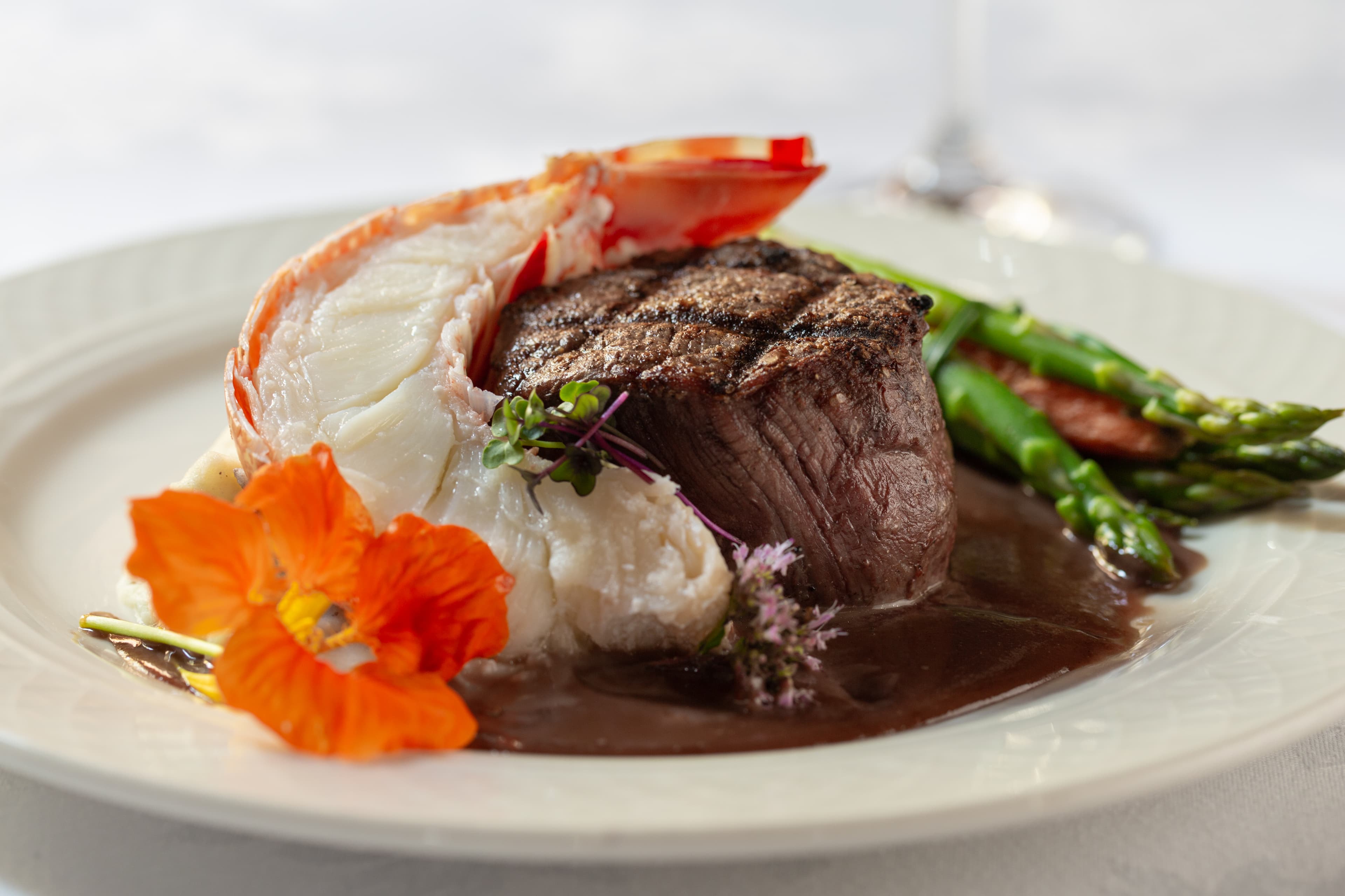 A gourmet dish featuring a grilled filet mignon topped with a lobster tail. The steak is served with steamed asparagus, mashed potatoes, and a rich brown sauce. An orange flower is placed on the plate for garnish.