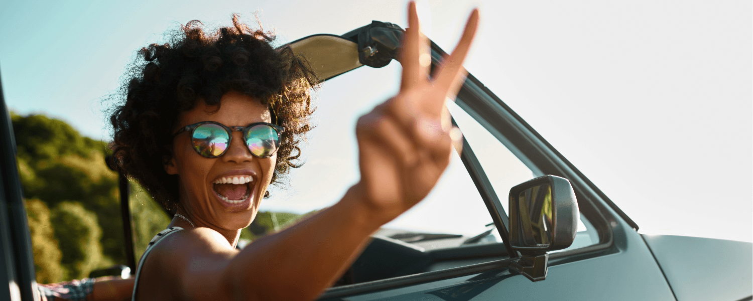 A smiling person in sunglasses flashes a peace sign from a car window.
