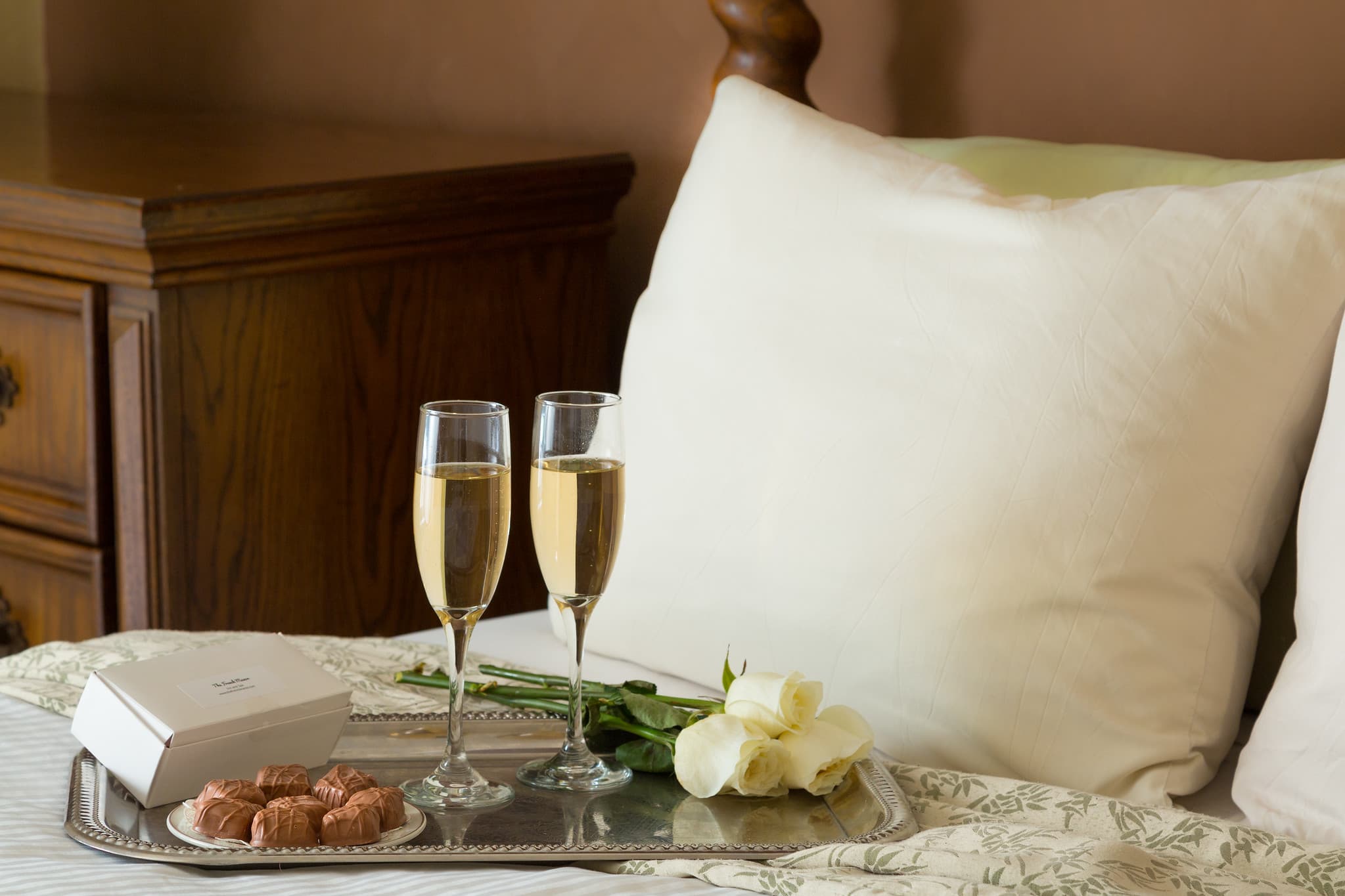 A tray with two champagne glasses, chocolate truffles, a white rose, and a small gift box is placed on a bed with decorative bedding.