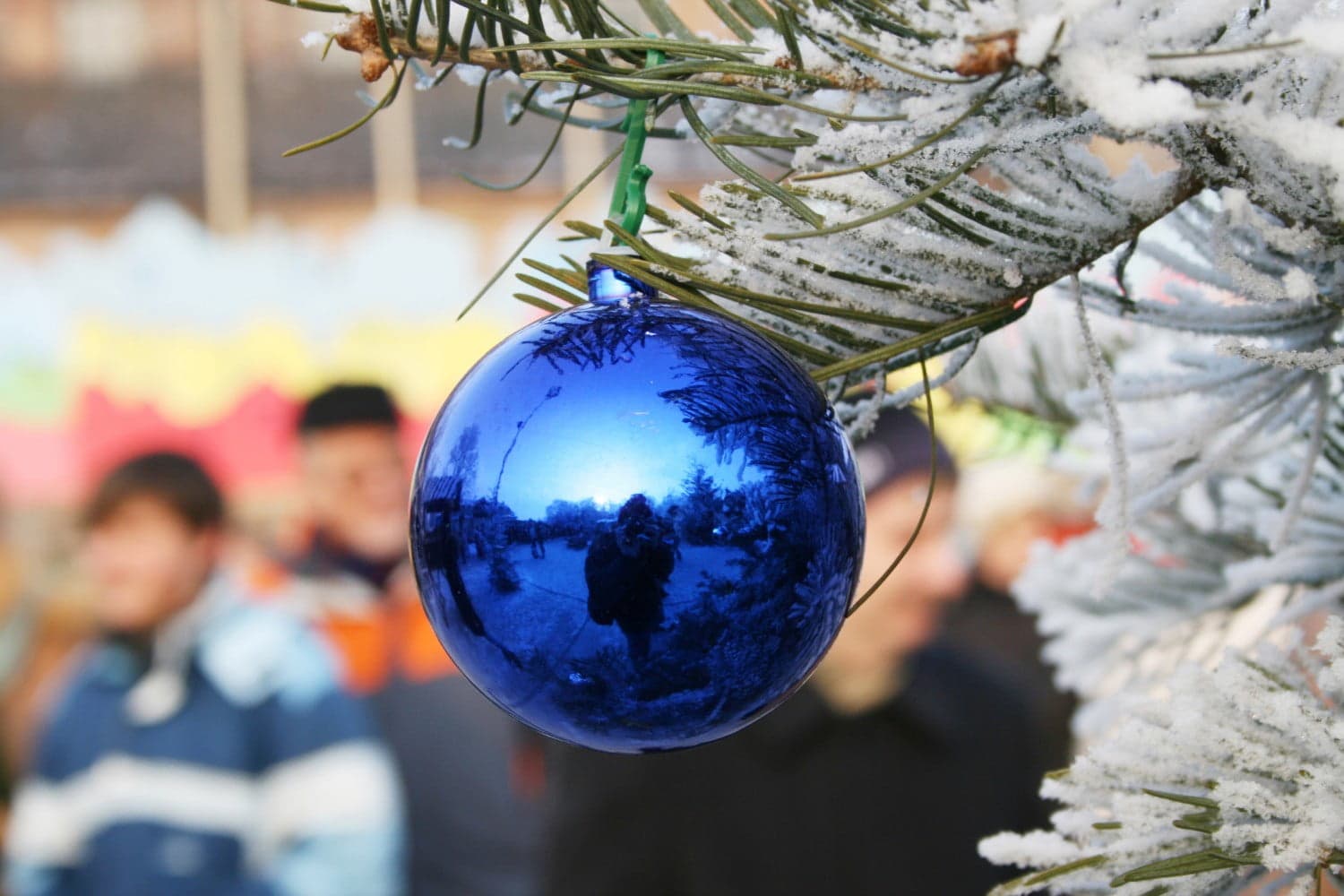 A reflective blue Christmas ornament hangs from a snowy pine branch, with blurred figures in the background.