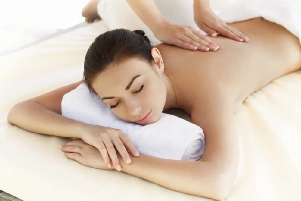 A woman laying face down on a massage table with her chin and face resting on a rolled towel, eyes closed, getting a back massage.