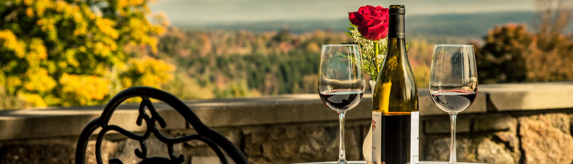 A bottle of wine with two glasses on a table, with a rose in a vase and a scenic view of the Pocono mountains in the background.