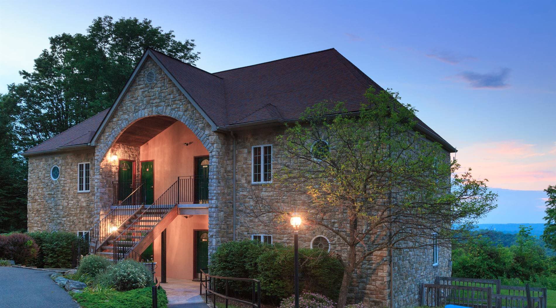 A stone building with a peaked roof and green doors, surrounded by trees and a sunset sky.