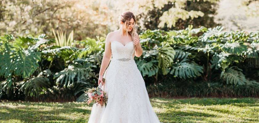 A bride in a white gown stands gracefully among lush greenery, holding a bouquet of flowers.