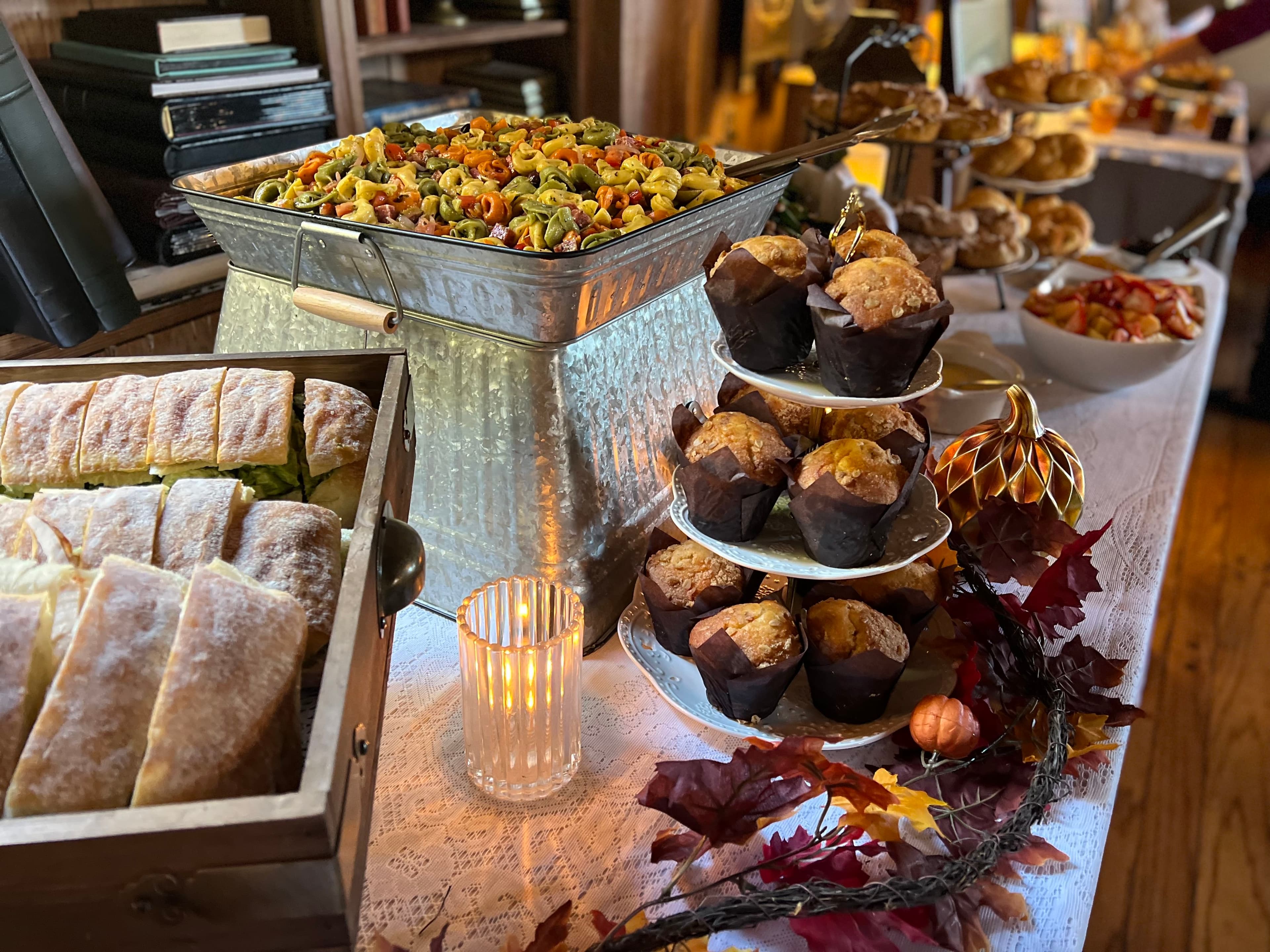 A table filled with food, including a metal container of pasta, a tiered stand of muffins, sandwiches in a wooden crate, and a bowl of fruit.