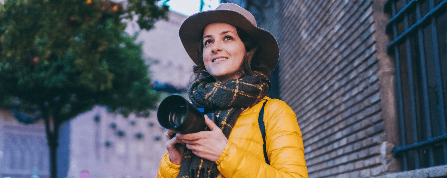 A woman wearing a yellow jacket and hat holds a camera while smiling outdoors.