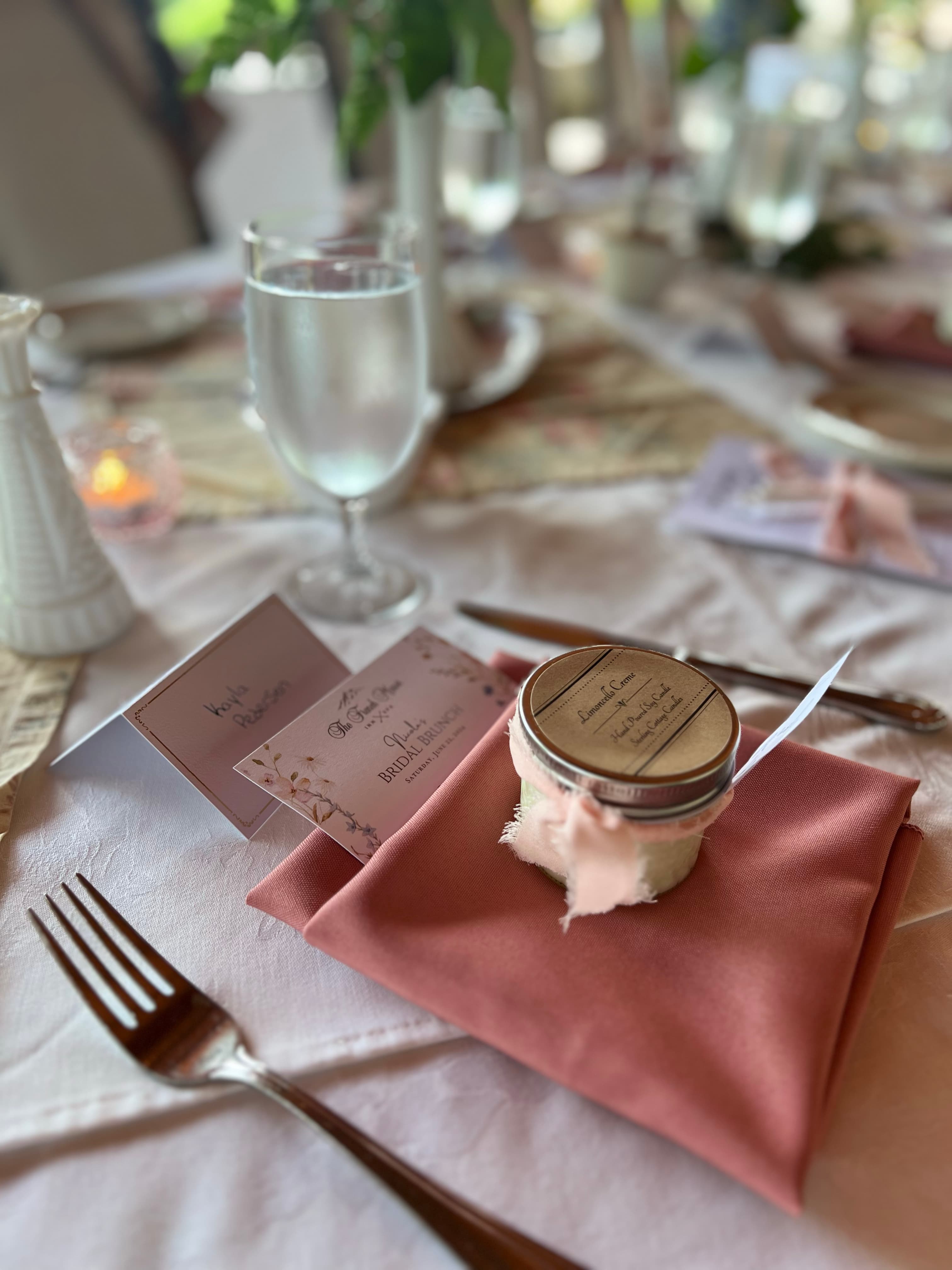 A place setting at a bridal brunch, featuring a peach-colored napkin, a glass of water, a personalized favor, and a place card with a floral design.