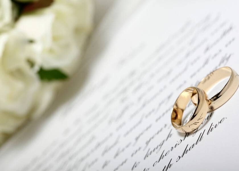 Two gold wedding rings resting on a wedding vow document beside a bouquet of flowers.
