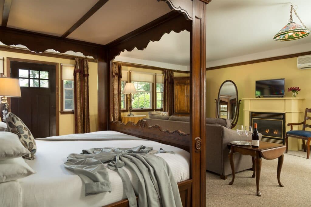 A cozy hotel room featuring a four-poster bed, a seating area, a fireplace, and windows with natural light.